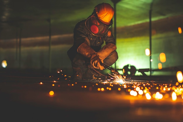 Chispa de corte de metal de trabajador masculino en la placa de acero del fondo del tanque con destello de luz de corte de cerca, use guantes protectores y máscara en el espacio confinado lateral.