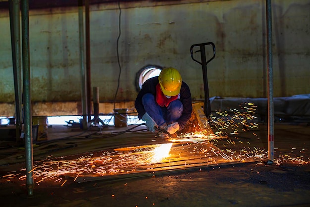 Chispa de corte de metal de trabajador masculino en la placa de acero del fondo del tanque con destello de luz de corte de cerca, use guantes protectores y máscara en el espacio confinado lateral.