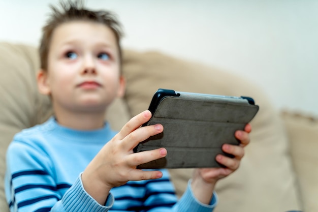 Chlid segurando o tablet pc enquanto está sentado em um sofá e olhando de lado as mãos do menino com um tablet sem fio no fundo desfocado