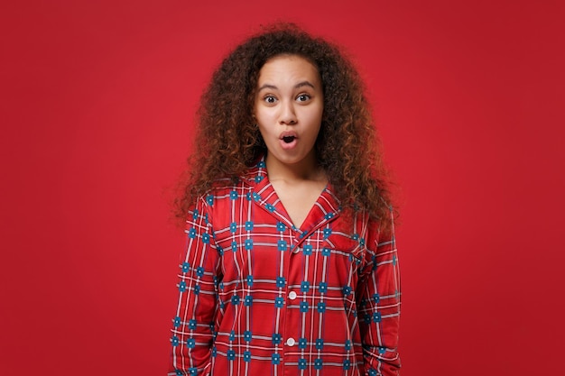 Chocada jovem afro-americana em homewear de pijama posando enquanto descansava em casa isolada em fundo vermelho no estúdio. Relaxe o conceito de estilo de vida de bom humor. Mock up espaço de cópia. Mantendo a boca aberta.