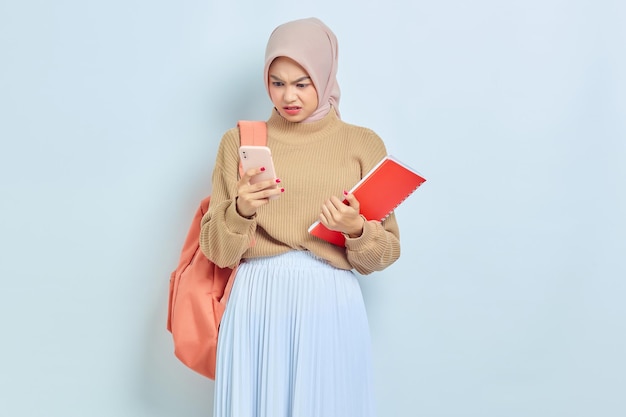 Chocada jovem estudante muçulmana asiática em suéter marrom com mochila segurando livro e celular isolado em fundo branco de volta ao conceito de escola