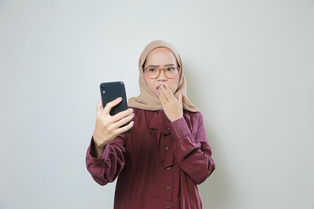 Chocada jovem muçulmana asiática usando óculos usando telefone celular isolado