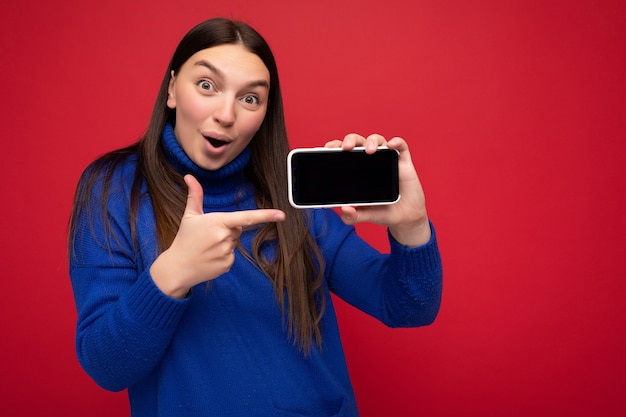 chocada muito feliz jovem mulher de parede vestindo um suéter azul casual isolado sobre o vermelho