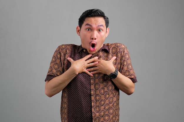 Chocado jovem asiático em camisa batik olhando para câmera com boca aberta isolada em fundo cinza