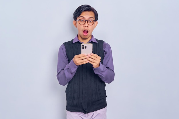 Chocado jovem asiático em camisa casual e colete usando um telefone celular recebendo ótimas notícias isoladas em fundo branco Conceito de estilo de vida de pessoas