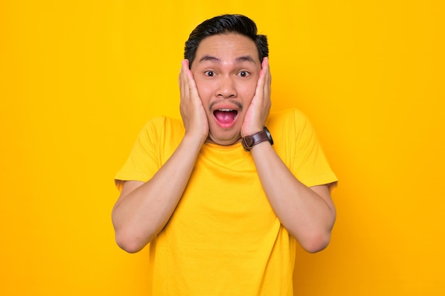 Chocado jovem asiático em camiseta casual com boca aberta e bochechas tocantes, olhando para a câmera com espanto isolado em fundo amarelo Conceito de estilo de vida de pessoas