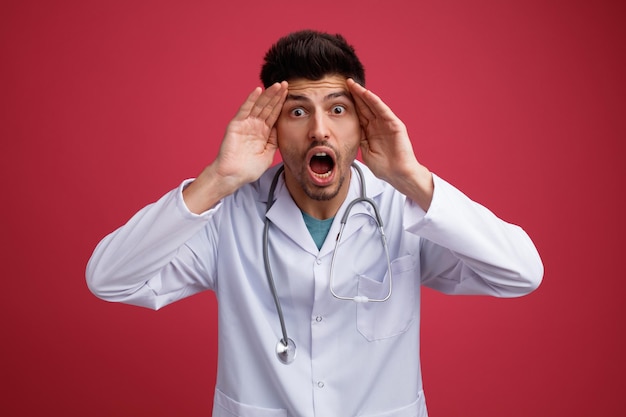 Chocado jovem médico masculino vestindo uniforme médico e estetoscópio olhando para a câmera mantendo as mãos na cabeça isolada em fundo vermelho