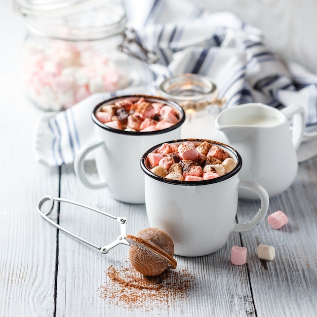 Chocolate caliente en dos tazas blancas esmaltadas con malvavisco sobre una mesa de madera clara.