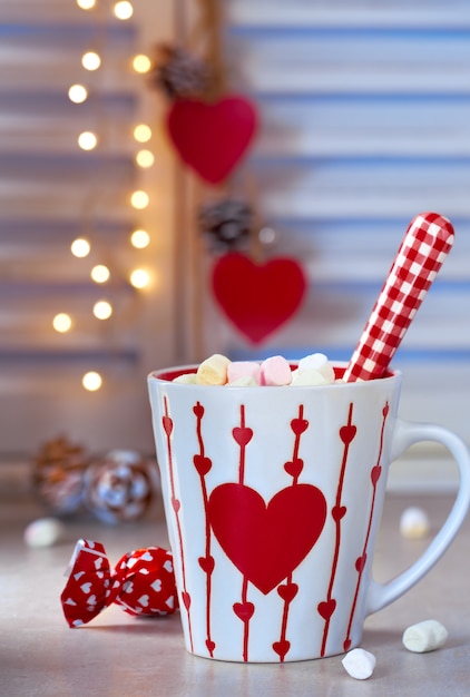 Chocolate caliente con malvaviscos, corazón rojo en la taza, pared de invierno con luces desenfocadas