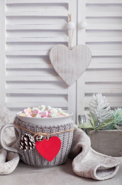 Chocolate caliente con malvaviscos, corazón rojo en la taza sobre la mesa con decoraciones de invierno