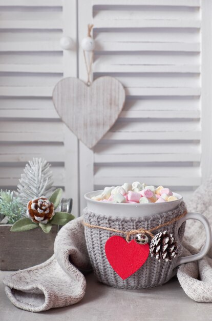 Chocolate caliente con malvaviscos, corazón rojo en la taza sobre la mesa con decoraciones de invierno