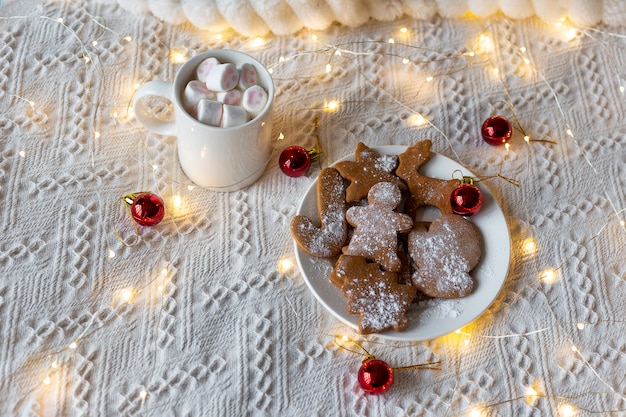 Chocolate caliente con malvaviscos y galletas de jengibre, guirnalda de luces festivas y juguetes de árbol de Navidad rojo en una cama blanca
