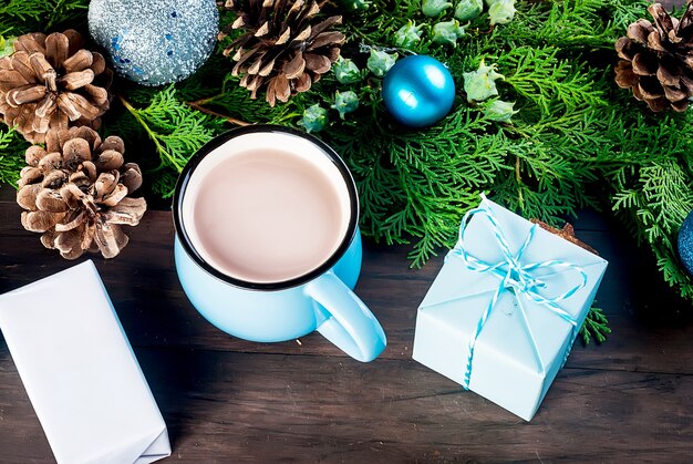 Chocolate caliente en taza azul sobre mesa de madera oscura con decoración navideña, regalos, piñas