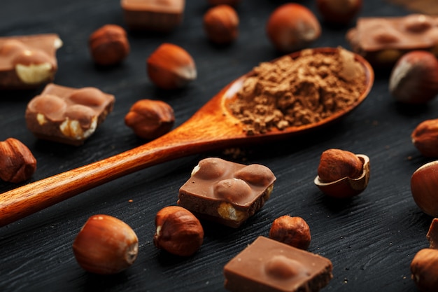 Foto chocolate com avelãs e uma colher de pau com cacau em um fundo escuro, rodeado de nozes com casca e descascado.