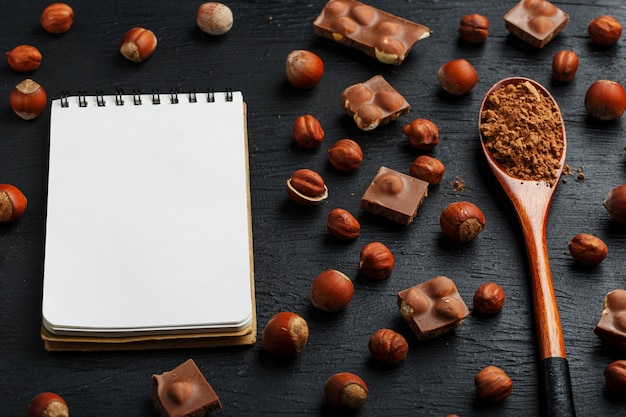 Foto chocolate com avelãs, um caderno com páginas em branco e uma colher de pau com cacau em um fundo escuro, rodeado de nozes com casca e descascado.