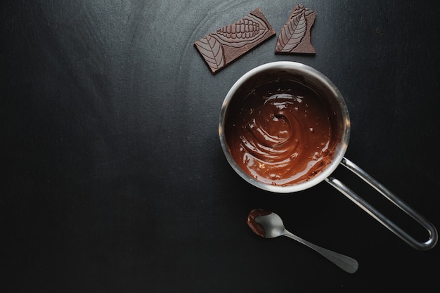 Chocolate derretido en una olla con trozos de chocolate alrededor de la mesa oscura. Vista superior.