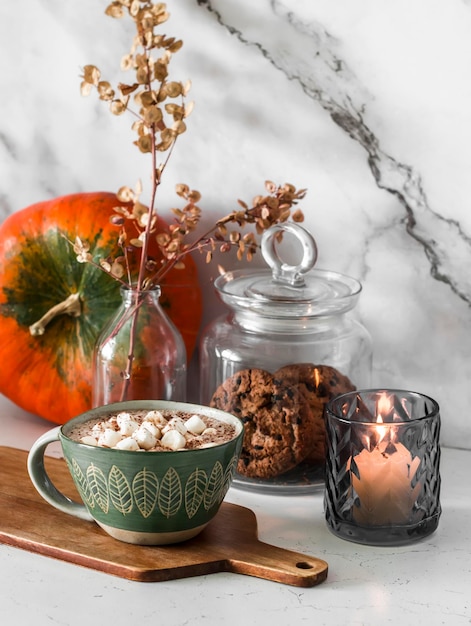 Chocolate quente com marshmallows e especiarias de torta de abóbora na mesa um pote de biscoitos uma vela acesa e uma abóbora interior de cozinha aconchegante