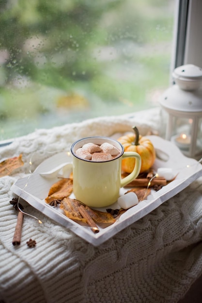 Chocolate quente com marshmallows em um copo amarelo. bandeja de madeira branca com decorações de xícara, abóbora e outono perto da janela com gotas de chuva