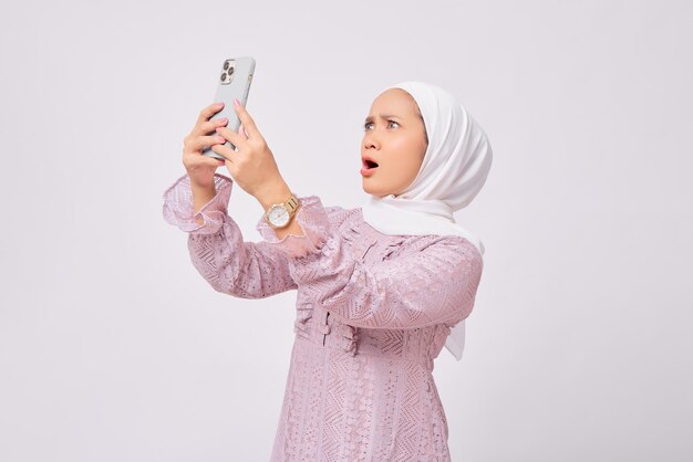 Chocou a bela jovem muçulmana asiática usando hijab e vestido roxo lendo notícias no celular e recebendo más notícias isoladas no fundo branco do estúdio