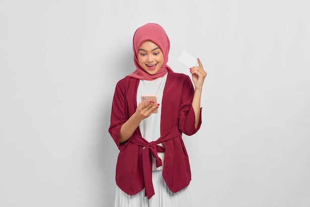 Chocou linda mulher asiática em uma camisa casual e hijab segurando um telefone celular, mostrando cartão de crédito, lendo boas notícias isoladas sobre fundo branco. Conceito de estilo de vida religioso de pessoas