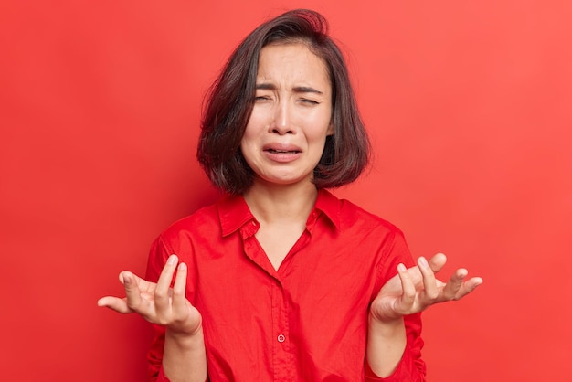 Chorando deprimida jovem asiática espalha palmas sente infeliz usa camisa enfrenta problemas fica gestos tristes de desespero isolado sobre fundo vermelho. Modelo feminino frustrado chora