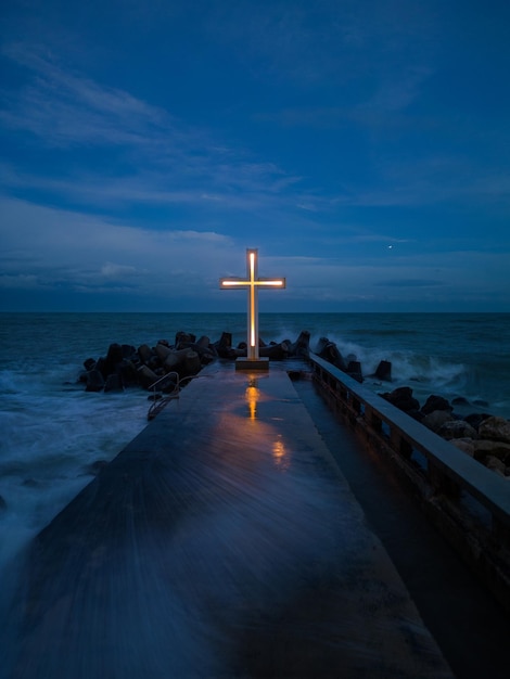 Christliches Kreuz, das nachts auf dem Pier im Meer oder Ozean mit dramatischem Himmel steht