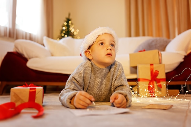 Christmas Child Escreva uma Carta para o Papai Noel com Chapéu de Natal Escrevendo uma Lista de Desejos de Presente ao lado da Árvore de Natal