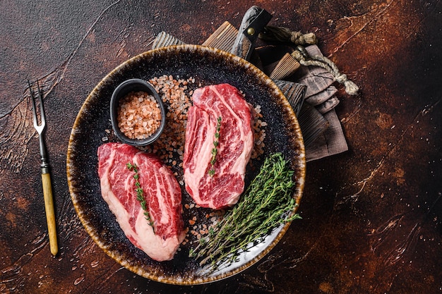 Chuck Eye Roll Rindersteaks, rohes Fleisch auf einem Teller mit Thymian und Kräutern. Dunkler Hintergrund. Ansicht von oben.