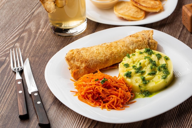 Chuleta de cerdo crujiente servida con puré de patata tibio espolvoreado con cebolla verde y ensalada de zanahoria en un plato blanco sobre una mesa de madera