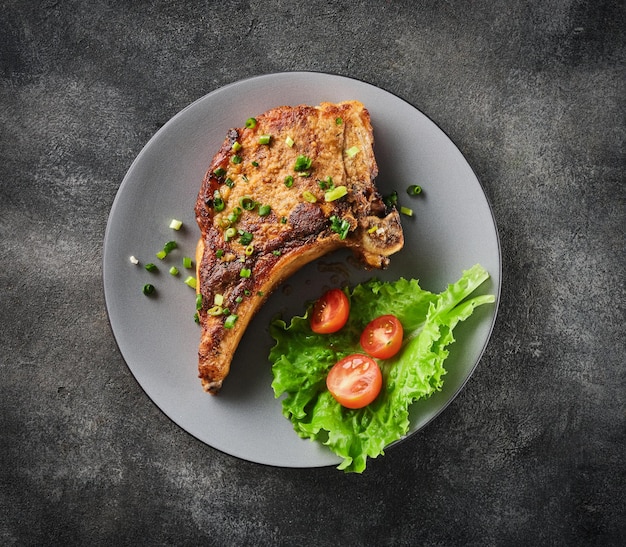 Chuleta de cerdo a la parrilla en un plato gris espolvoreado con cebollas verdes con ensalada y tomates cherry