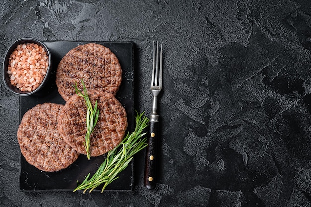 Chuleta de hamburguesa de ternera para hamburguesa a la parrilla en barbacoa en tablero de mármol con romero Fondo negro Vista superior Espacio de copia