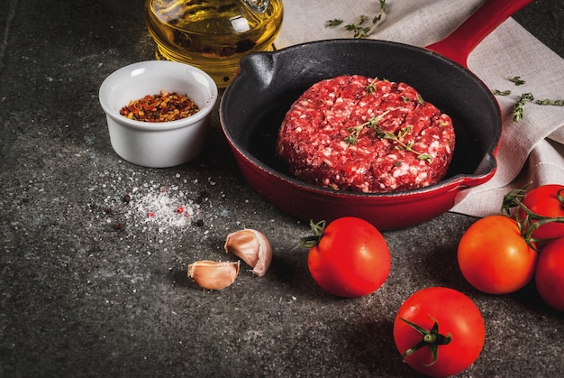 Chuletas de carne cruda orgánica de carne de res con especias, tomillo, tomates, aceite de oliva en una sartén sobre una superficie negra