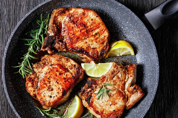 Chuletas de cerdo asadas con ramitas de romero y rodajas de limón en una sartén sobre una mesa de madera, vista horizontal desde arriba, endecha plana