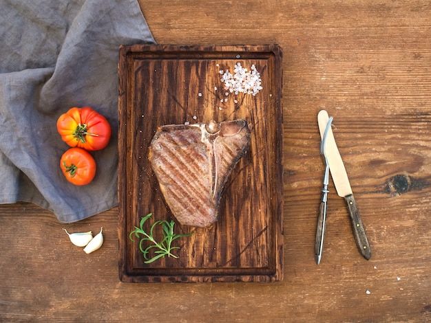 Chuletón de carne cocida sobre tabla de servir con dientes de ajo, tomates, romero y especias sobre una rústica mesa de madera