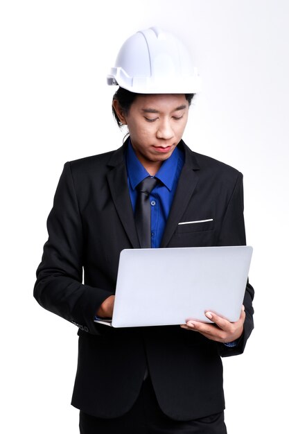 Chupito de estudio aislado de ingeniero industrial asiático profesional exitoso capataz masculino en traje formal negro y casco de seguridad permanente escribiendo informe en equipo portátil sobre fondo blanco.