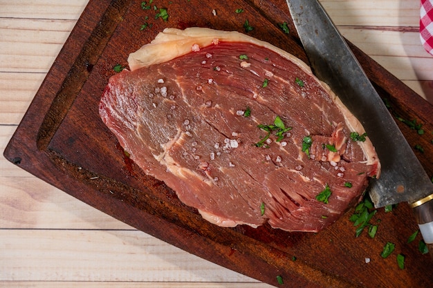 Churrasco de cuadril o bistec crudo sobre una tabla de cortar de madera rústica con hierbas. Vista superior.