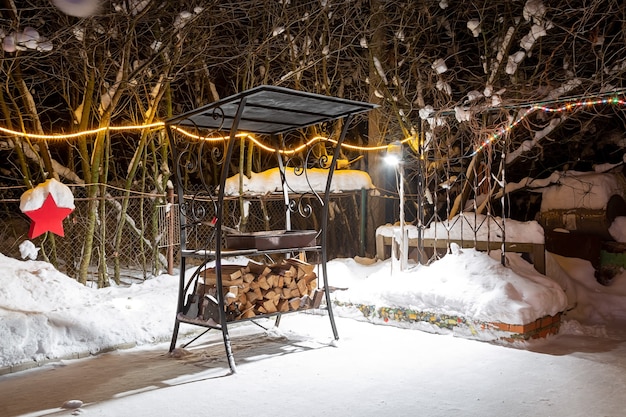 Churrasco perto de casa no inverno. Noite, guirlandas estão queimando, está nevando. Preparação para assar carne. Eles colocam a lenha colhida sob a grelha.