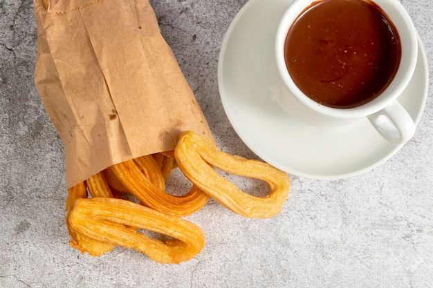 Churros en bolsa de papel con taza de chocolate caliente, desayuno típico español, sobre fondo gris piedra