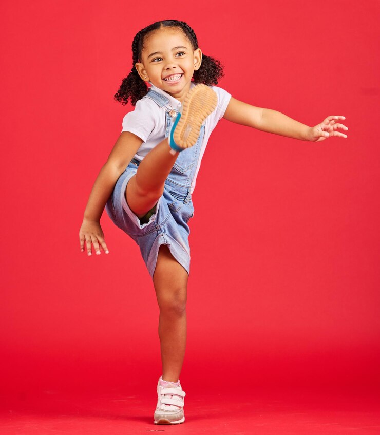 Foto chute feliz e garota animada em um estúdio com uma roupa de criança elegante e casual sorriso de felicidade e criança pequena com roupas legais chutando a perna com emoção isolada por um fundo vermelho