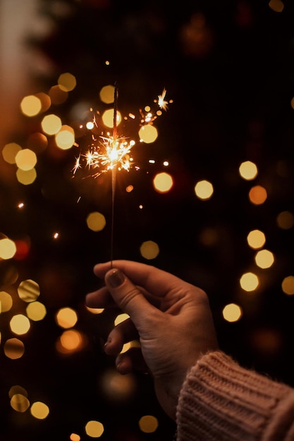 Foto chuveiro ardente na mão no fundo das luzes douradas do bokeh no quarto escuro festivo feliz ano novo mão segurando fogos de artifício na árvore de natal com iluminação dourada