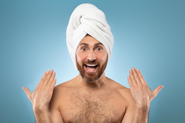 Chuveiro matinal e rotina de beleza Engraçado homem barbudo caucasiano de meia-idade posando com toalha na cabeça sobre fundo azul