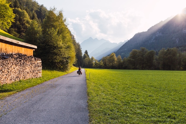 Ciclista con alforjas pedaleando en un carril bici en los Alpes austríacos con las montañas y el sol entre las montañas