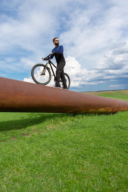 Ciclista barbudo en una bicicleta de montaña en un tubo oxidado, hierba verde, cielo azul