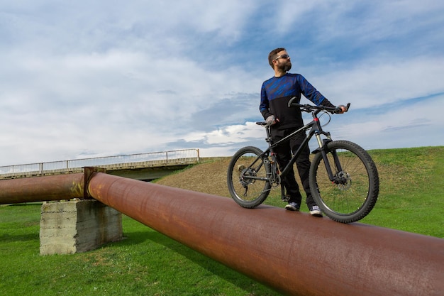 Ciclista barbudo en una bicicleta de montaña en un tubo oxidado, hierba verde, cielo azul