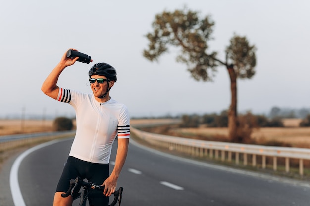 Ciclista barbudo cansado en casco negro y gafas salpicaduras de agua de botella deportiva mientras está de pie en la carretera con su bicicleta