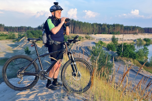El ciclista con la bicicleta y los prismáticos en la mano se encuentra en una colina de arena en la luz del sol