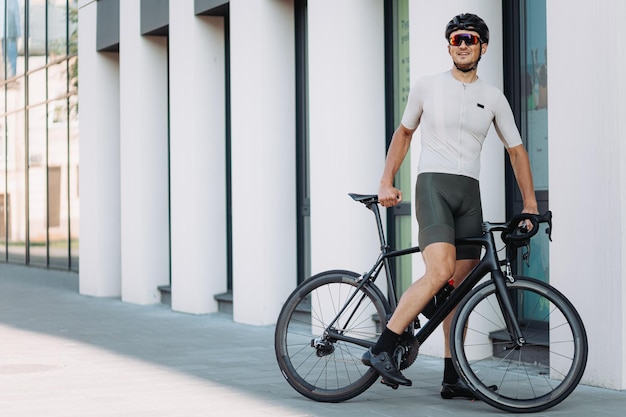 Ciclista caucásico en casco de seguridad y ropa activa de pie cerca de un edificio moderno con su bicicleta negra Hombre guapo descansando después de entrenar durante el tiempo libre al aire libre