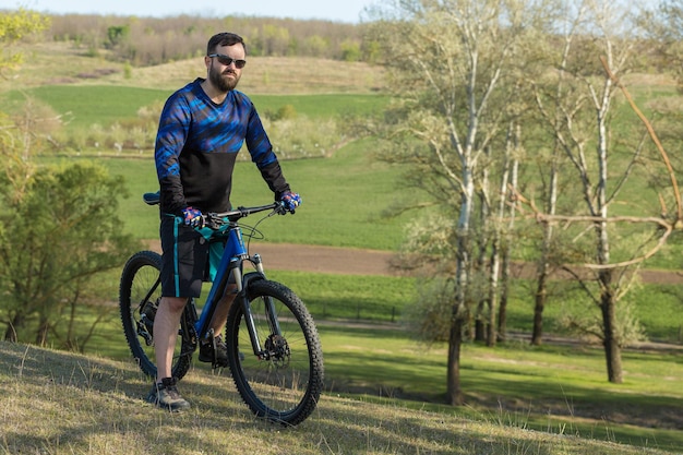 Ciclista de bermuda e malha em uma moderna bicicleta rígida de carbono com um garfo de suspensão a ar em um penhasco contra o fundo de uma floresta verde fresca de primavera