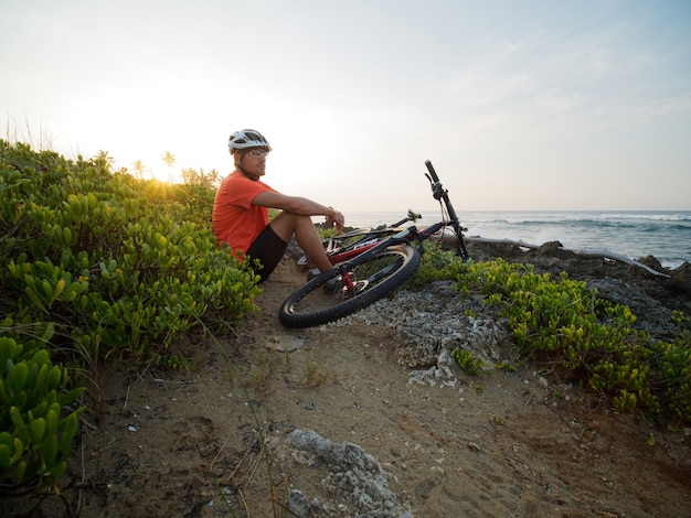 Ciclista de homem usando capacete branco e assento de camiseta laranja com uma mountain bike, na costa do oceano. Praia rochosa.