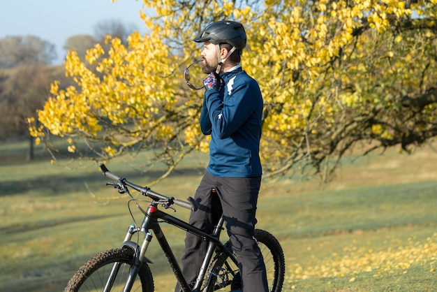 Ciclista em calças e jaqueta de lã em uma moderna bicicleta hardtail de carbono com um garfo de suspensão a ar passeios offroad O cara está descansando em um banco no parque outono